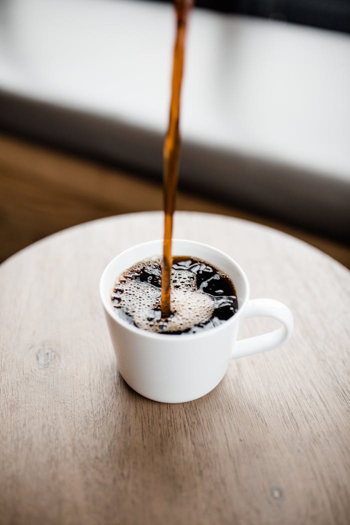 brand-03 Steaming black coffee being poured into a white ceramic mug on a wooden table.